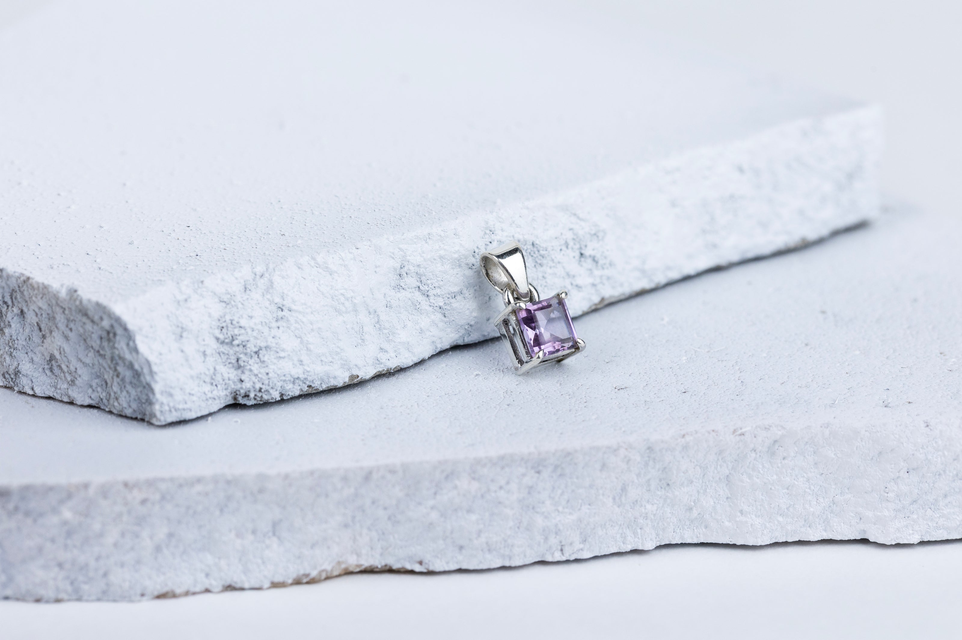 A square-cut amethyst gemstone pendant mounted on a silver setting, displayed against a grey and white background.