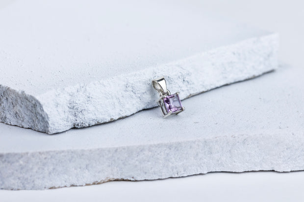 A square-cut amethyst gemstone pendant mounted on a silver setting, displayed against a grey and white background.