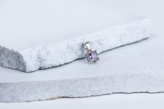 A square-cut amethyst gemstone pendant mounted on a silver setting, displayed against a grey and white background.