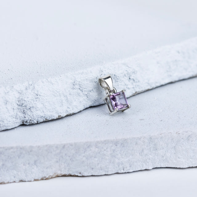 A square-cut amethyst gemstone pendant mounted on a silver setting, displayed against a grey and white background.